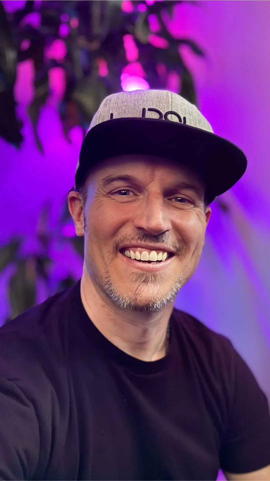 Joshua Taylor – Founder of The Taylor Vibe™ | Digital Strategist & Kingdom Entrepreneur Smiling portrait of Joshua Taylor in a black shirt and branded hat, with a purple-lit background and greenery.